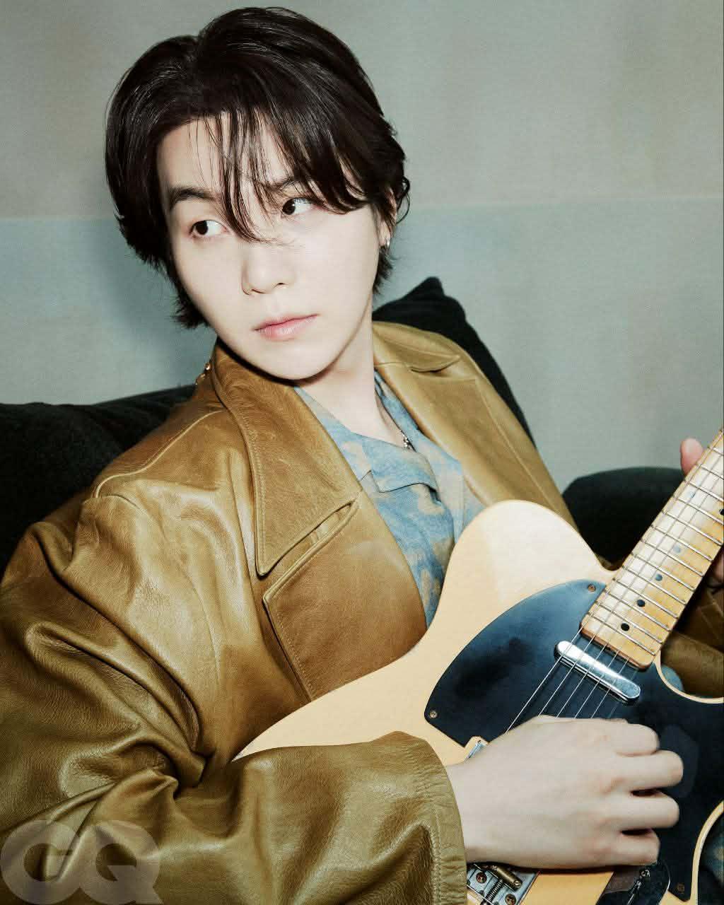 Suga of BTS seated on a sofa with a guitar on his lap. He is wearing a brown leather jacket and a blue printed shirt, and he is looking to the left.