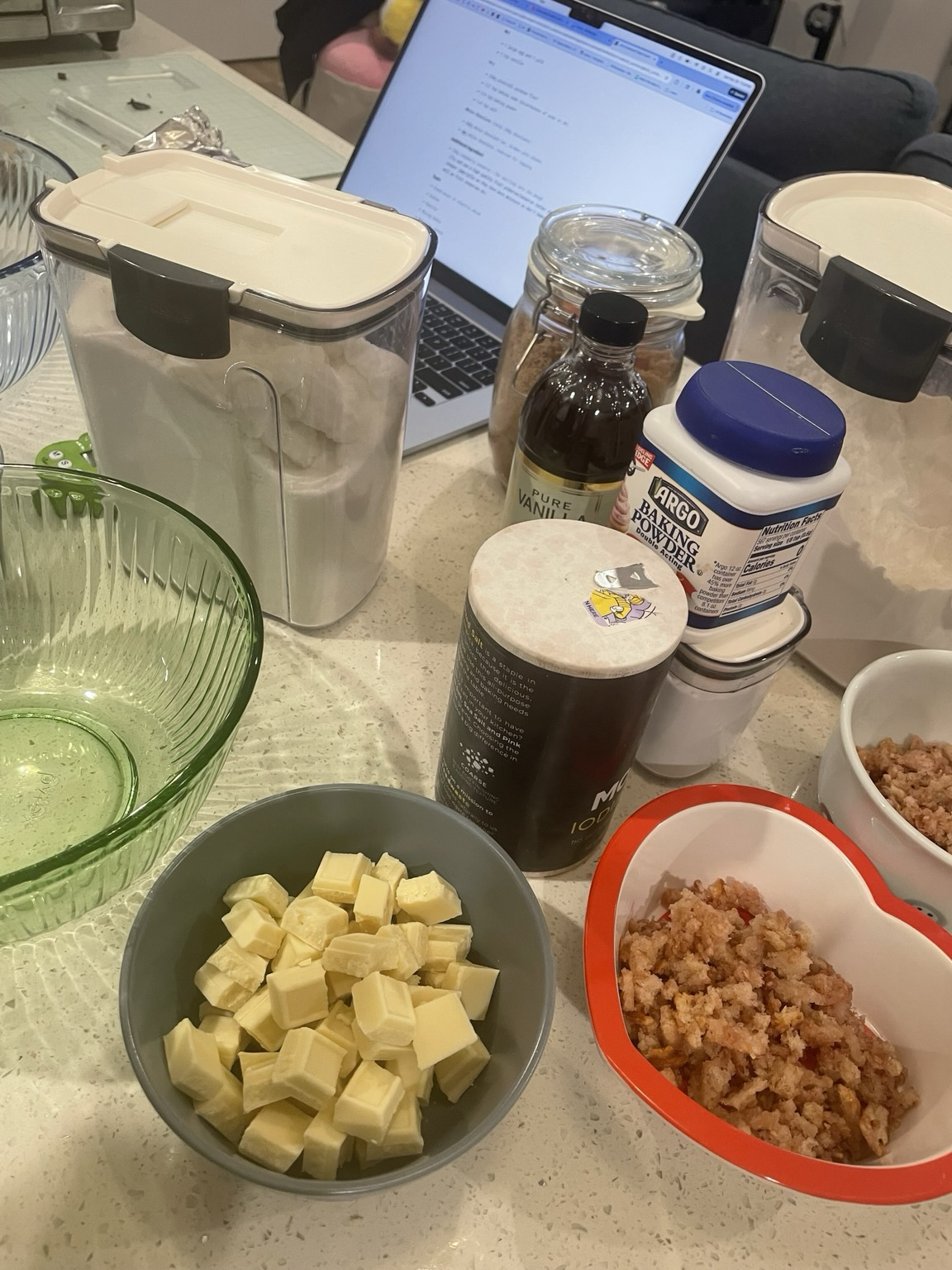 Various baking ingredients and accessories - a green empty bowl, granulated sugar, salt, baking powder, baking soda, vanilla extract, dark brown sugar, flour, toasted crumbs, and and white chocolate chunks