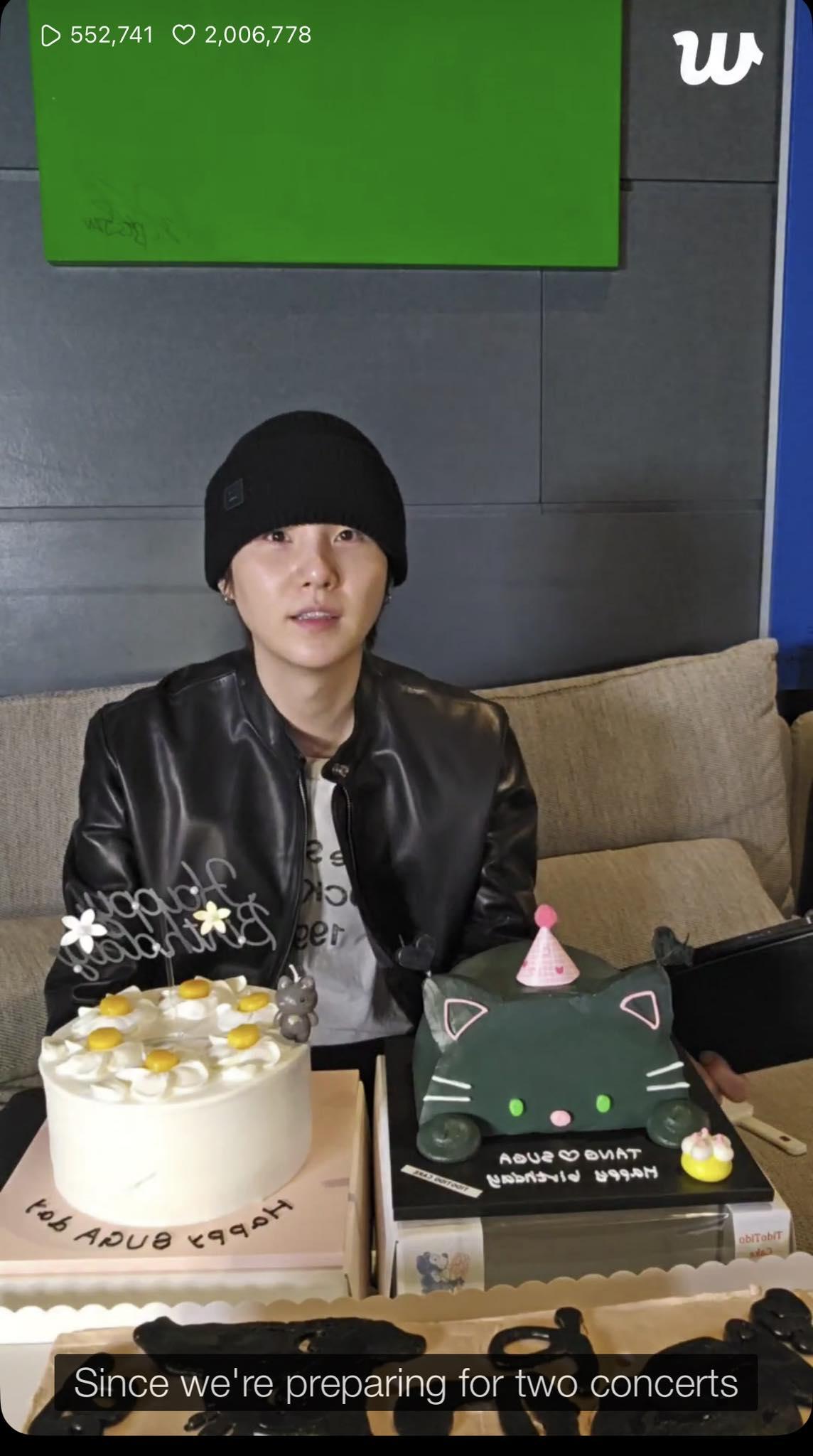 Suga of BTS in front of his two birthday cakes. He is wearing a black leather jacket and a black beanie. The cakes are a white cream cake and a black cat fondant cake.
